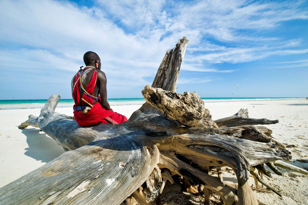 A Maasai wearing traditional clothes is sitting on the beach.